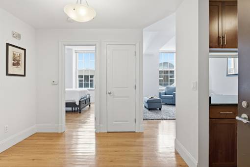 A white hallway with a bedroom and a kitchen visible through the door.
