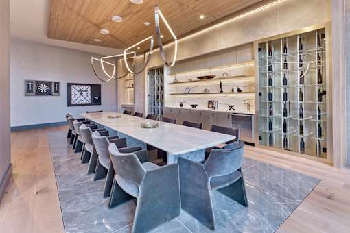 A long white table with grey chairs is in front of a shelf with glass doors.