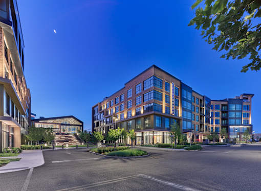 A modern multi-story building with a glass facade is surrounded by trees and other buildings.