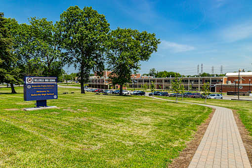 A blue sign with white text is in the foreground of a grassy area with a brick path leading to a parking lot.