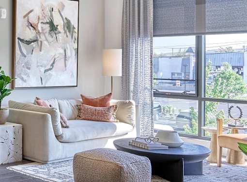 A living room with a white couch, a grey ottoman, a table with a lamp and a painting on the wall.