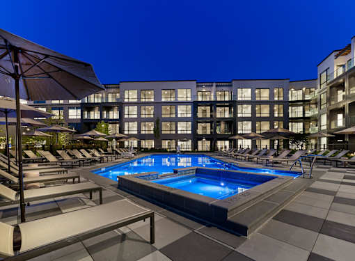 A large swimming pool surrounded by lounge chairs and umbrellas in front of a modern building.