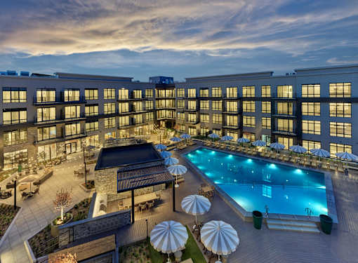 A large swimming pool surrounded by lounge chairs and umbrellas is the centerpiece of a resort-style courtyard in a luxury apartment rental community