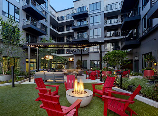 A fire pit sits in the middle of a grassy area surrounded by red chairs.