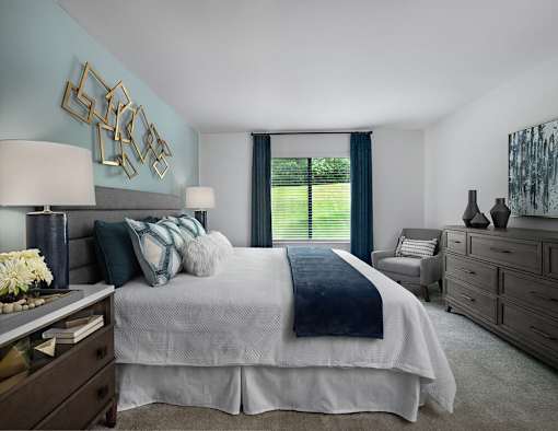 Spacious bedroom at River Oaks at Rochester Hills Apartments with king bed, accent wall, dresser and window seat.