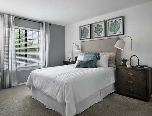 Bright bedroom at River Oaks at Rochester Hills Apartments with white bed linens, two nightstands, lamps, and window blinds.