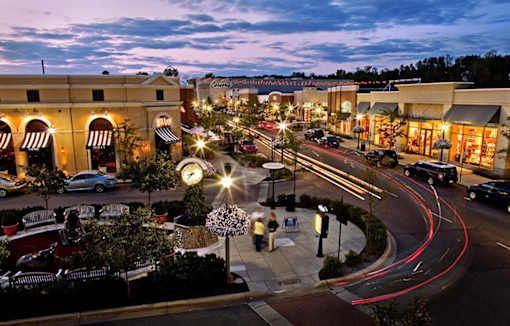 Dusk view of lit plaza and storefronts near River Oaks at Rochester Hills Apartments.