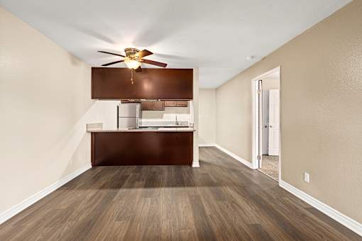 A room with a ceiling fan and a wooden floor leading to a kitchen area.