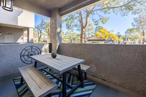 A wooden table and bench are on a patio with a wall and trees in the background.