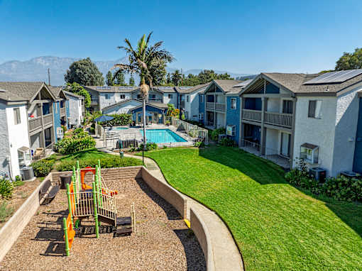 A playground with a slide and swings is surrounded by a grassy area and townhomes.