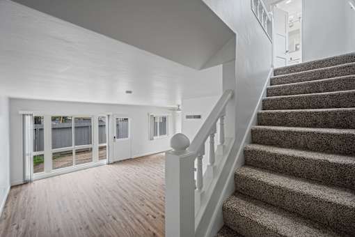 A staircase with a carpeted runner and white balusters leads to a brightly lit room with white walls and a wooden floor at Hillsdale Garden Apartments, San Mateo, CA 94403