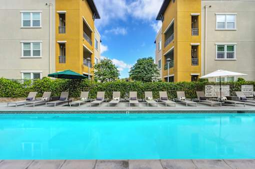 A swimming pool surrounded by lounge chairs and umbrellas in front of apartment buildings.