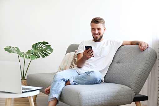 A man is sitting on a grey couch with a laptop and a phone in his hands.