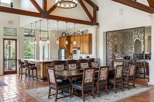 A large wooden dining table with chairs is set up in a room with a kitchen in the background.