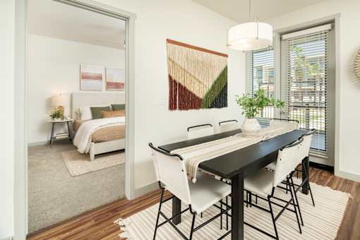 A dining table with four chairs is in the foreground of a room with a bed in the background.