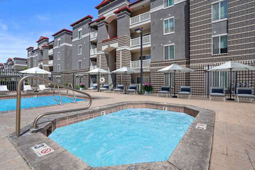 A swimming pool surrounded by lounge chairs and umbrellas in front of a multi-story apartment building.