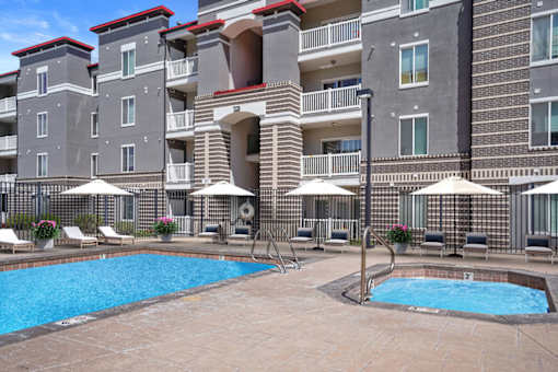 A large swimming pool surrounded by lounge chairs and umbrellas in front of a multi-story apartment building.