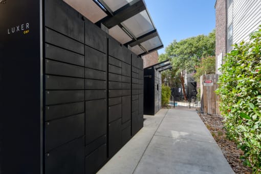 a black garage door opens to a long sidewalk with a bush on the side