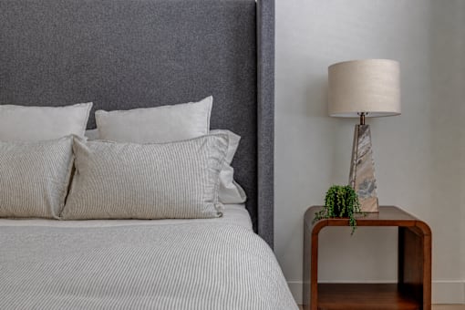 A bed with white pillows and a grey headboard is next to a wooden side table with a lamp and a plant.