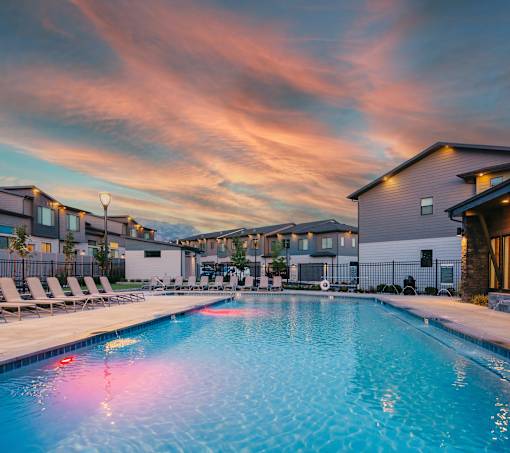 A swimming pool surrounded by lounge chairs and buildings under a colorful sky.