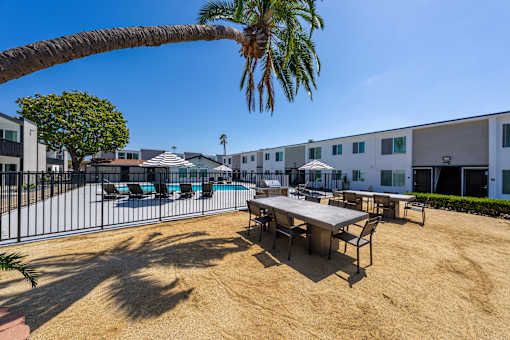 A sandy area with a table and chairs and a pool in the background.