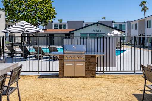 A BBQ grill is in the middle of a sandy area with a pool and umbrellas in the background.
