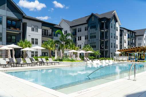 A swimming pool surrounded by lounge chairs and umbrellas in front of a row of apartment buildings.
