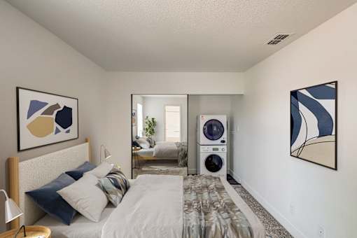 A bedroom with a bed, pillows, a nightstand, a washing machine, and a painting on the wall.