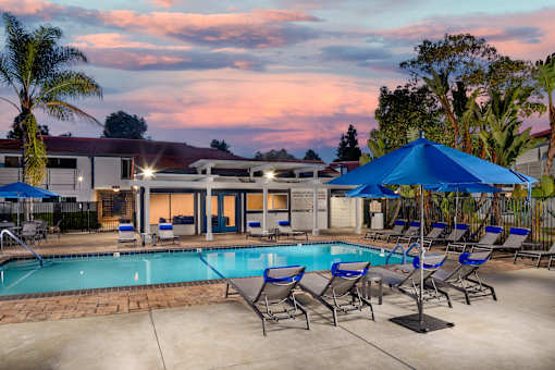 A poolside area with lounge chairs and umbrellas is set against a sunset sky.