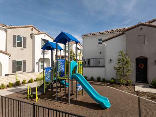 A playground with a blue slide and yellow safety barriers in front of a white house at San Carlo Townhomes Apartments, Highland, CA, 92346