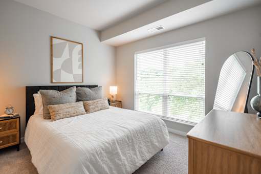 A bedroom with a large bed, a nightstand, a lamp, and a window with blinds.
