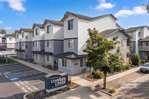 A sunny day at the Logger Creek Apartments with clear blue skies and a few cars parked in the lot.