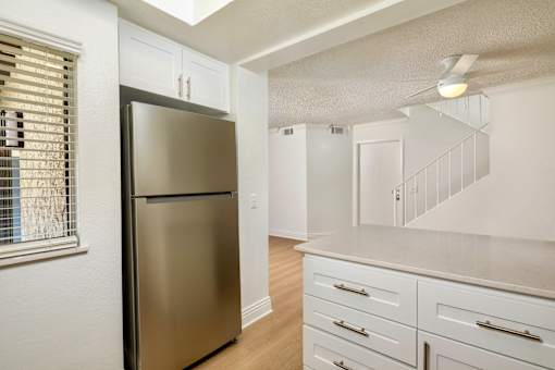 A stainless steel refrigerator in a kitchen with white cabinets and a window with blinds.