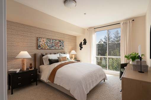 A bedroom with a large bed, a lamp, a painting, and a window with a view of trees.