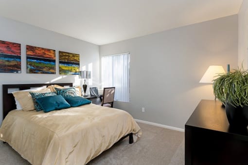 A bedroom with a bed, nightstand, lamp, and a plant at The Casas Apartments, San Diego, CA