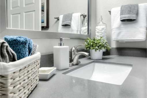 A white sink with a basket of towels and a potted plant on a grey countertop.