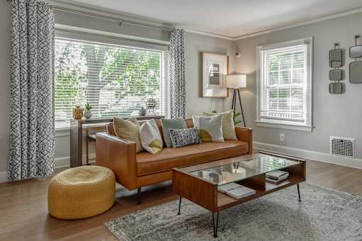 A living room with a brown couch, a glass coffee table, and a yellow pouf.