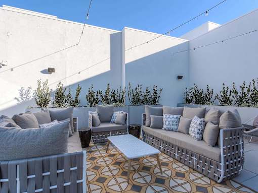 A patio with a white couch, grey pillows, and a patterned rug.