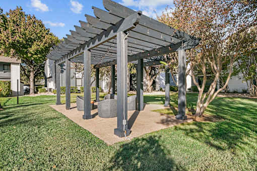 A large grey pergola with a slatted roof is situated in a grassy area.