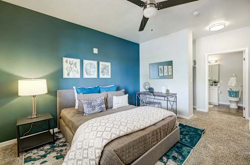 A bedroom with a bed, a nightstand with a lamp, a ceiling fan, and a chair.