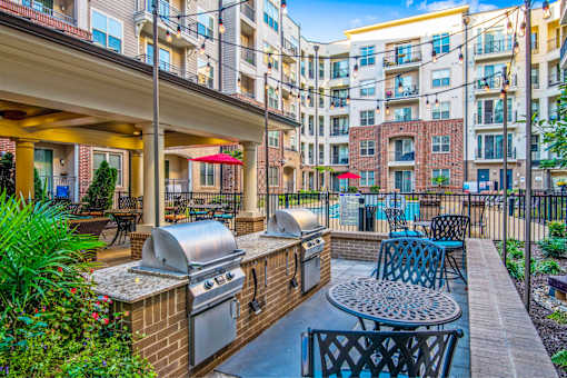A patio with a grill and chairs is surrounded by a brick wall and buildings.