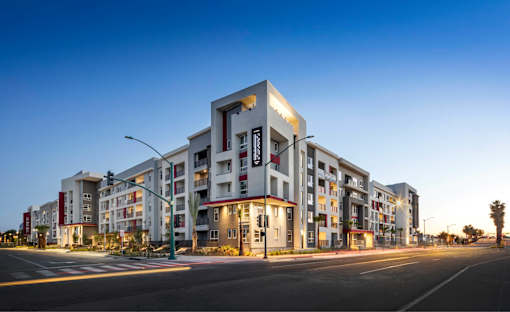 A modern apartment building with multiple floors and balconies is illuminated by streetlights at dusk
