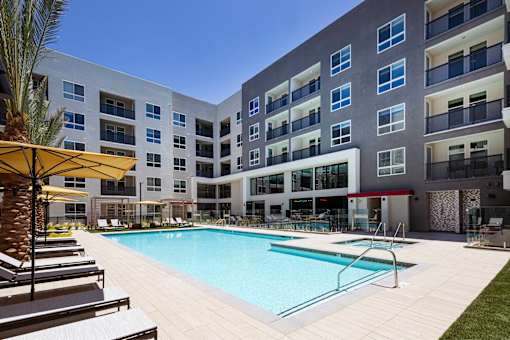 A large swimming pool is surrounded by sun loungers and umbrellas in front of a modern apartment building.