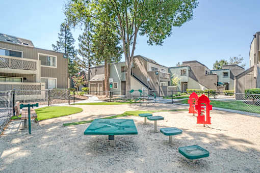 A playground with a green table and red chairs in the foreground and apartment buildings in the background.