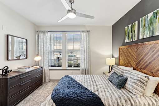 An interior view of a bedroom with a bed, a front end table with drawers, two large windows with curtains, a mirror, and a ceiling fan at Parkstone at Knightdale apartments for rent in Knightdale, NC.