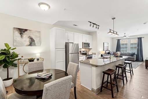 An interior view of a modern kitchen with a kitchen island countertop, a dining area with a dining table and chairs, and views of a living room with two large windows, a ceiling fan, and a grey couch at Parkstone at Knightdale apartments for rent in Knightdale, NC.