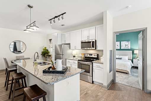 An interior view of a modern kitchen with a kitchen island countertop, barstool chairs, stainless steel appliances, and views of a bedroom at Parkstone at Knightdale apartments for rent in Knightdale, NC.