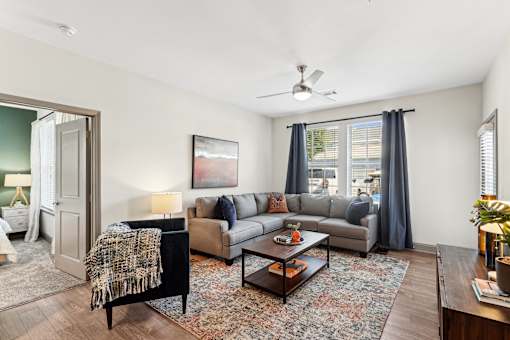 An interior view of a living room with a grey couch, a black chair, a coffee table, and a ceiling fan, with partial views of a bedroom at Parkstone at Knightdale apartments for rent in Knightdale, NC.