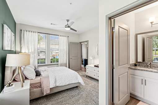 An interior view of a bedroom with a bed, a side table with a lamp on it, two large windows with curtains, a closet door, and a bathroom with a mirror and a granite countertop at Parkstone at Knightdale apartments for rent in Knightdale, NC.