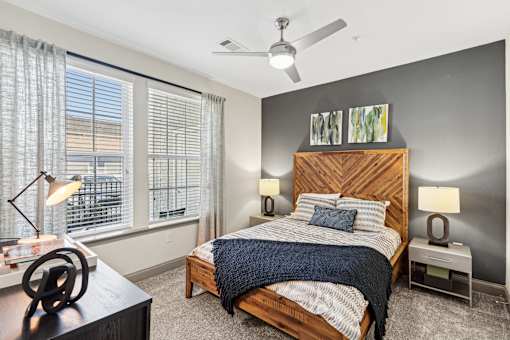 An interior view of a bedroom with a bed, two side tables with lamps on them, a ceiling fan, a front end table, and two large windows with curtains at Parkstone at Knightdale apartments for rent in Knightdale, NC.
